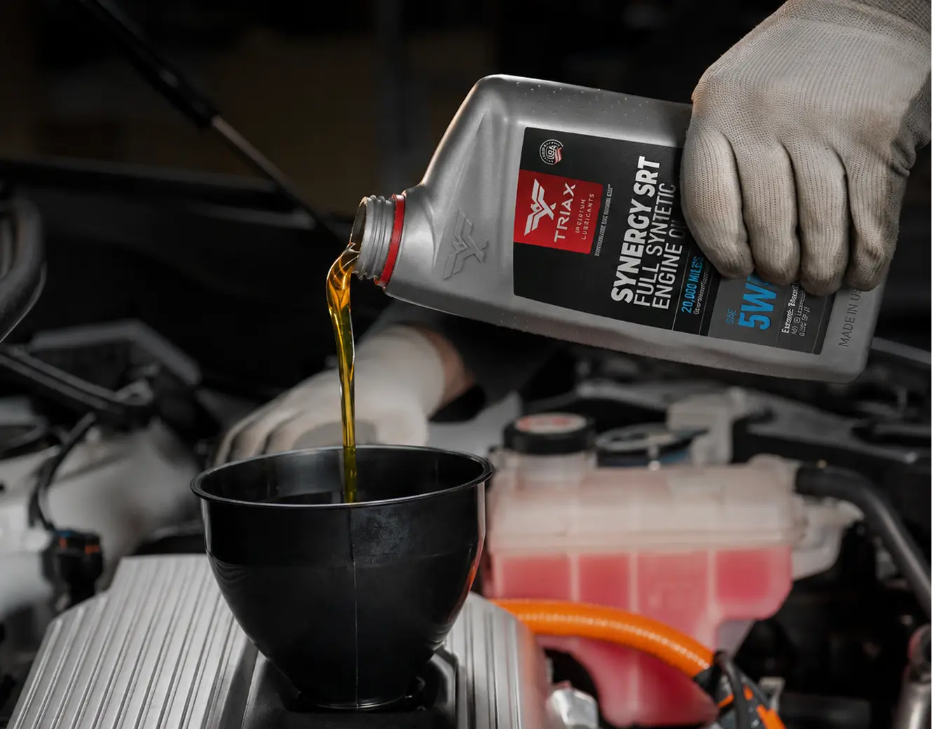 A gloved hand holds a bottle of TRIAX Synergy SRT Full Synthetic Engine Oil, pouring golden oil into a black funnel placed in an engine bay.