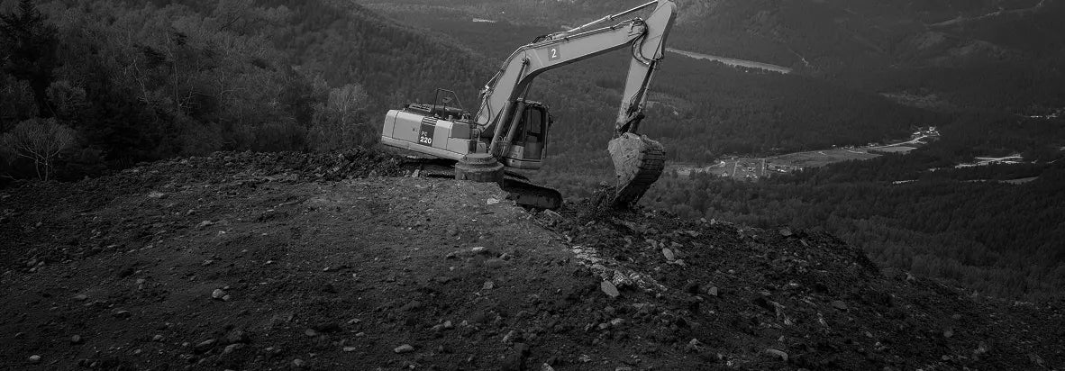 Black and white photograph of an excavator positioned on a steep, rocky hillside with a forested valley in the background.