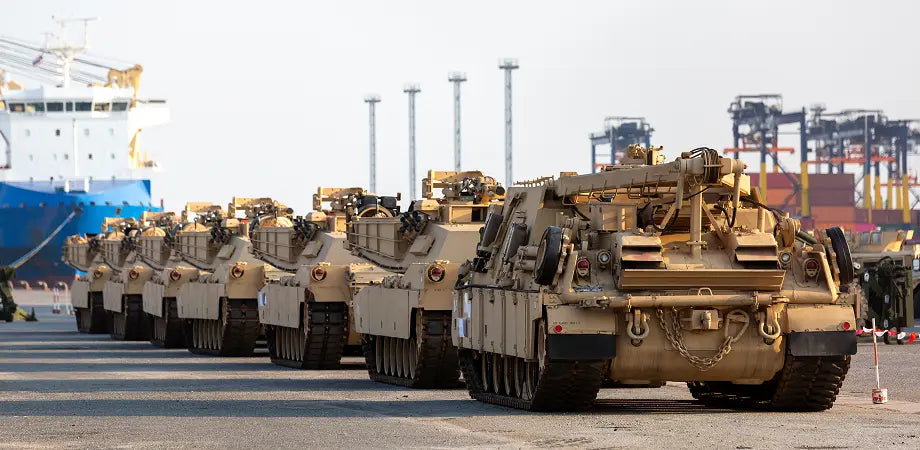 Row of military tanks on a road with industrial buildings in the background